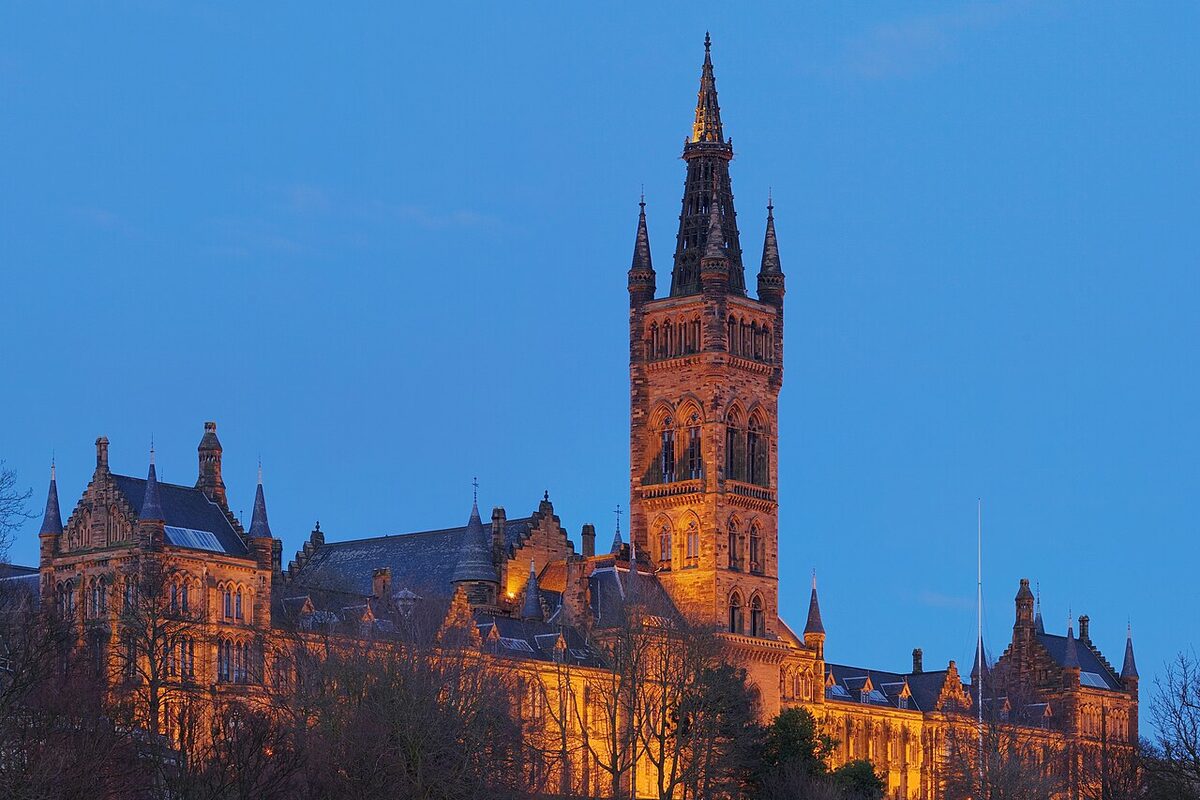 The University of Glasgow's Gilbert Scott Building, floodlit at dusk against a blue sky